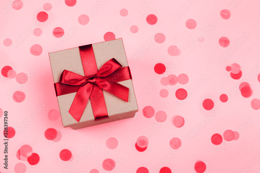Present box with red bow on pastel pink background with multicolored confetti. Flat lay style.