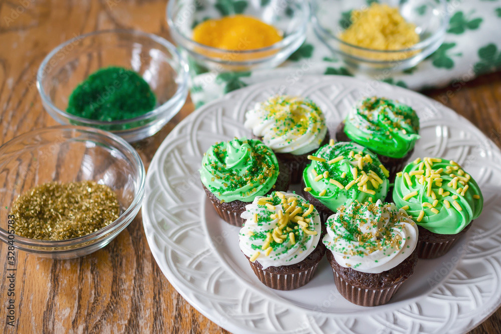 A plate of green and white frosted chocolate miniature cupcakes decorated with gold, yellow, and green assorted sprinkles. Sprinkles blurred in background on rough wooden table. From above. A plate of green and white frosted chocolate miniature cupcakes decorated with gold, yellow, and green assorted sprinkles. Sprinkles blurred in background on rough wooden table. From above.