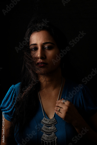 Close up face portrait of a beautiful Indian Bengali brunette woman in light and shadow before a black copy space background wearing a blue top. Indian lifestyle and fashion portrait