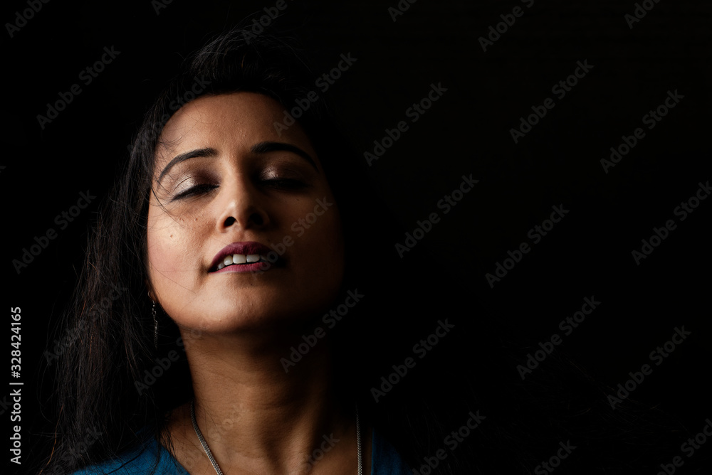 Close up face portrait of a beautiful Indian Bengali brunette woman in light and shadow before a black copy space background wearing a blue top. Indian lifestyle and fashion  portrait