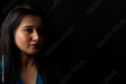 Close up face portrait of a beautiful Indian Bengali brunette woman in light and shadow before a black copy space background wearing a blue top. Indian lifestyle and fashion  portrait