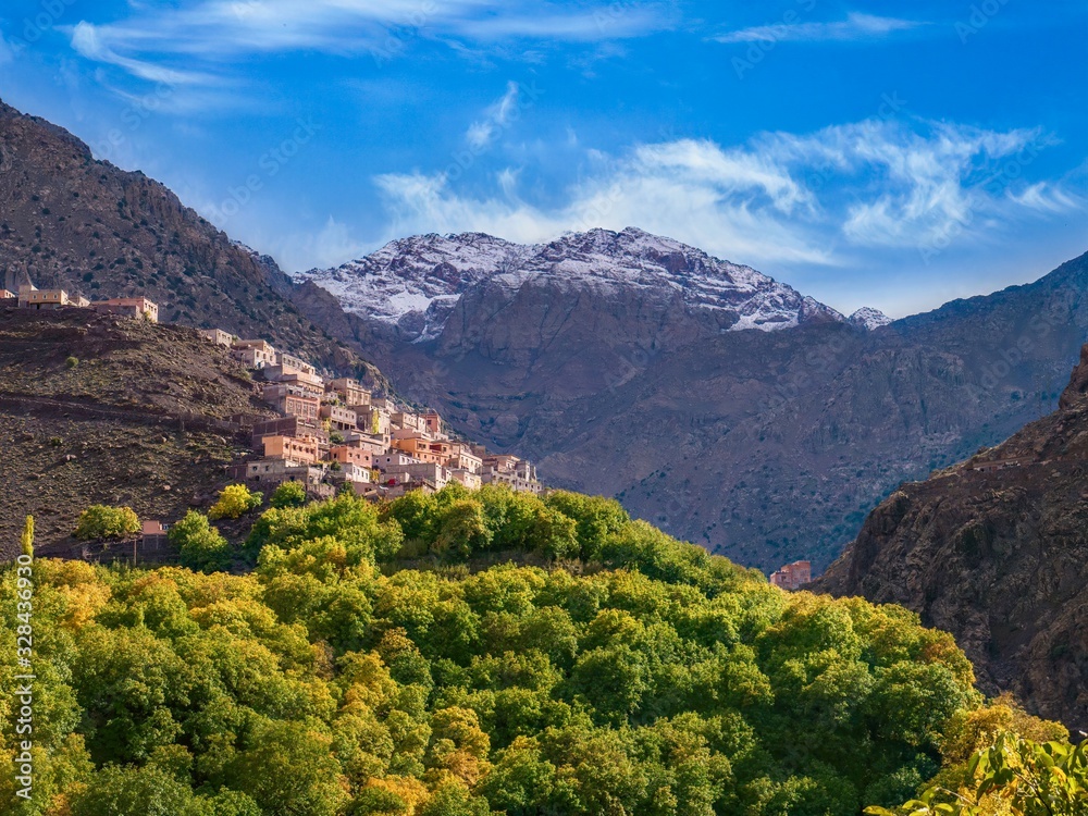 In the High Atlas mountains of Morocco, a Berber village is perched on a steep hillside, with snowcapped Jbel Toubkal, the tallest mountain in North Africa, in the background.