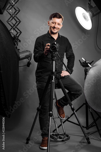 Happy man photographer with camera on a studio equipment background. Portrait of a smiling male model in black fashion clothes taking photos in the studio. Self portrait