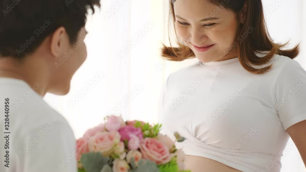 Asian pregnant wife is happy with her unborn baby. Husband giving her a bouquet for their wedding anniversary and kiss on her stomach to talk with their child. Asian pregnant wife is happy with her unborn baby. Husband giving her a bouquet for their wedding anniversary and kiss on her stomach to talk with their child.