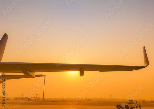 Closeup airplane wing with golden sunrise sky. Plane parked at airfield of airport. Morning flight. Summer travel concept. Pushback tractor support with airport staff at taxiways. Aviation business.