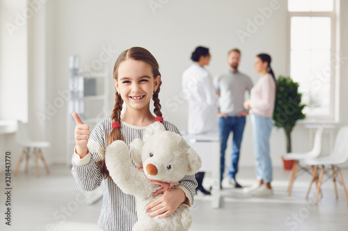 Fototapeta Naklejka Na Ścianę i Meble -  Girl with a toy stands against the background of the doctor and parents standing in the clinic office.