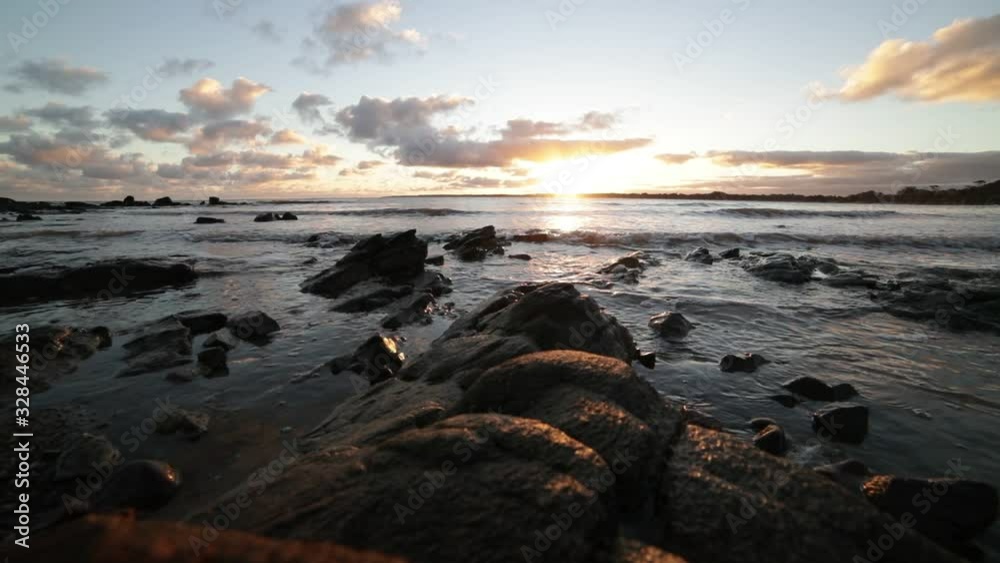 Slow motion of beach at golden hour. Foreground of rocks at shoreline while waves brake at the sand. Golden reflection over sea surface. Sunset at the horizon. Piriapolis, Uruguay