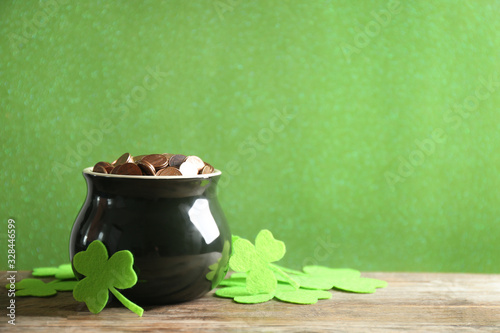 Pot of gold coins and clover leaves on wooden table against green background, space for text. St. Patrick's Day celebration