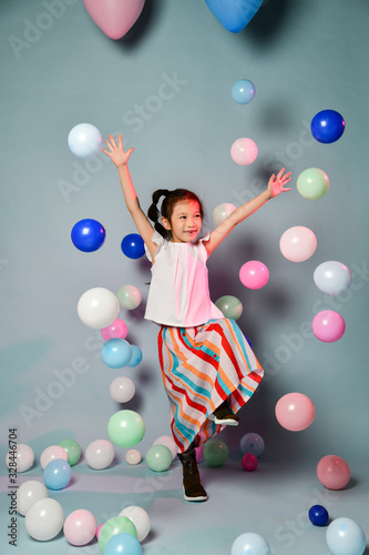 Happy asian kid girl in stylish striped maxi skirt has fun jumps skipping dances surrounded by colorful air balloons