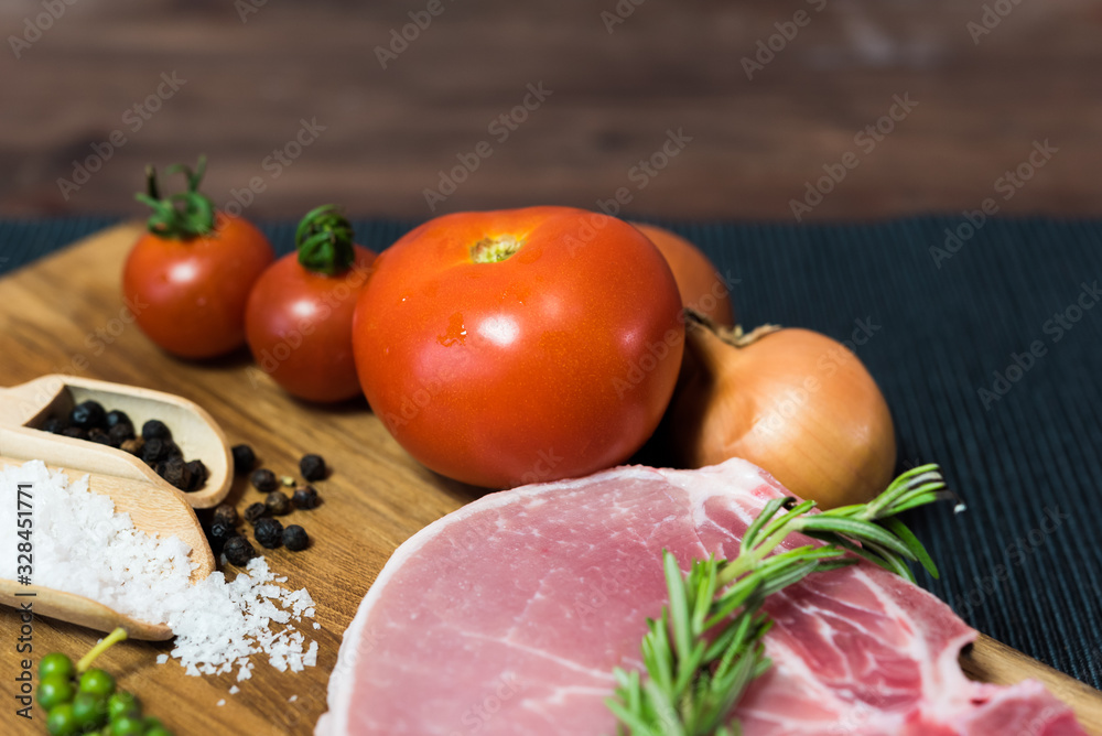 Raw pork chop meat on wood plate with vegetables, spices, salt and black pepper in dark black cloth table. Focus on tomato ingredient Raw pork chop meat on wood plate with vegetables, spices, salt and black pepper in dark black cloth table. Focus on tomato ingredient