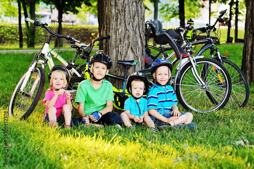a group of small preschool children in Bicycle safety helmets smile sitting on the fresh green grass in the Park against the background of bicycles and trees.