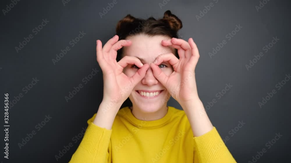 Cheerful funny young woman having fun making glasses shape with hands, does funny face with eyes wide open, dressed in yellow sweater, isolated on grey background. People emotions concept Cheerful funny young woman having fun making glasses shape with hands, does funny face with eyes wide open, dressed in yellow sweater, isolated on grey background. People emotions concept