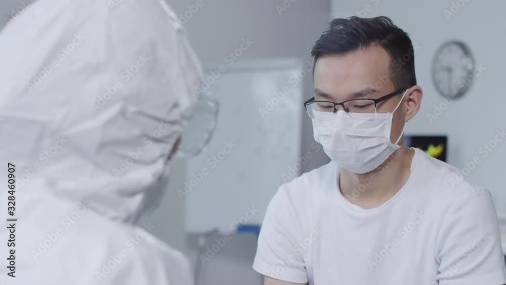 Over the shoulder shot of unrecognizable doctor in disposable coveralls, goggles and gloves checking temperature of Asian male patient in face mask Over the shoulder shot of unrecognizable doctor in disposable coveralls, goggles and gloves checking temperature of Asian male patient in face mask