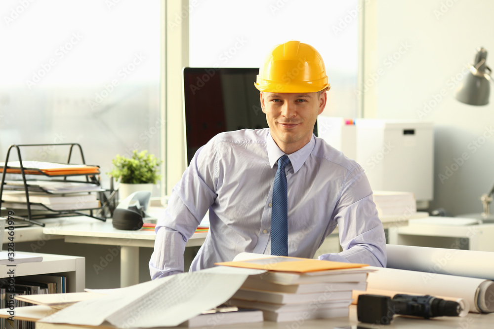Smiling Civil Engineer Working on House Sketch. Glad Architect Sitting with Layout Plan of Rooms at Working Table. Man Wearing Shirt and Yellow Helm. Reconstruction Service Worker Looking at Camera Smiling Civil Engineer Working on House Sketch. Glad Architect Sitting with Layout Plan of Rooms at Working Table. Man Wearing Shirt and Yellow Helm. Reconstruction Service Worker Looking at Camera