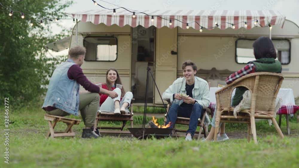 Young guy in casual outfit holding stick with marshmallow and telling story to diverse friends while sitting near fire during trip in nature Young guy in casual outfit holding stick with marshmallow and telling story to diverse friends while sitting near fire during trip in nature