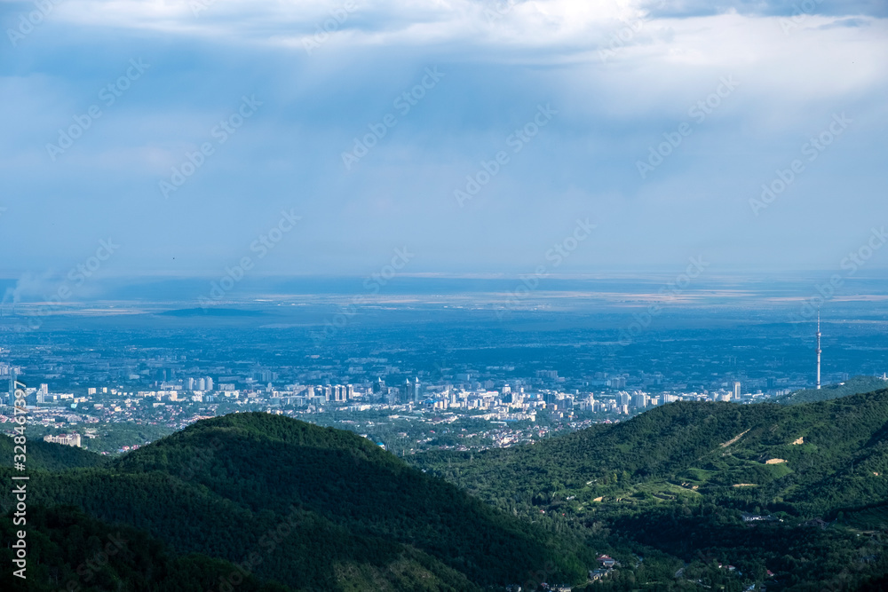 Beautiful view to Almaty city and foothills with storm clouds captured from mountains. Green almaty mountains foothills clouds in beautiful style on blue background.