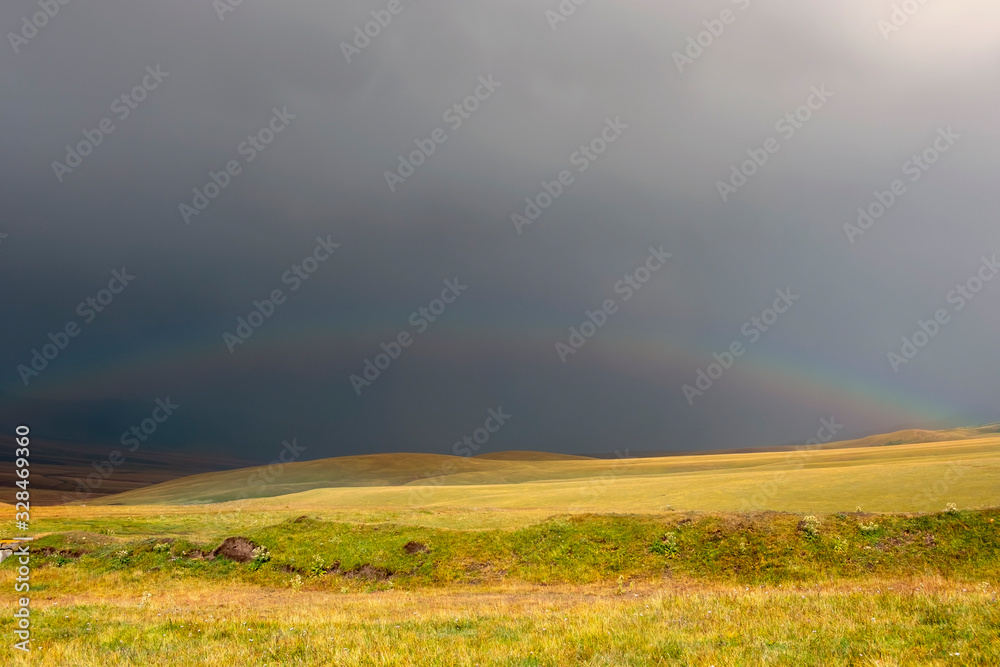 Beutiful rainbow above green meadow after heavy rain with rain clouds and mountains background. Summer season. Fresh green grass. Travel background. Assy plateau near Almaty city in Kazakhstan.