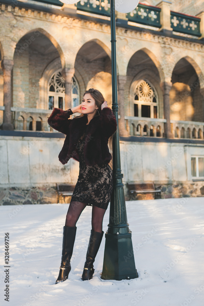 Fashion model with red lips under street light posing and watching a sun. Sunlight on the background. Snow around.