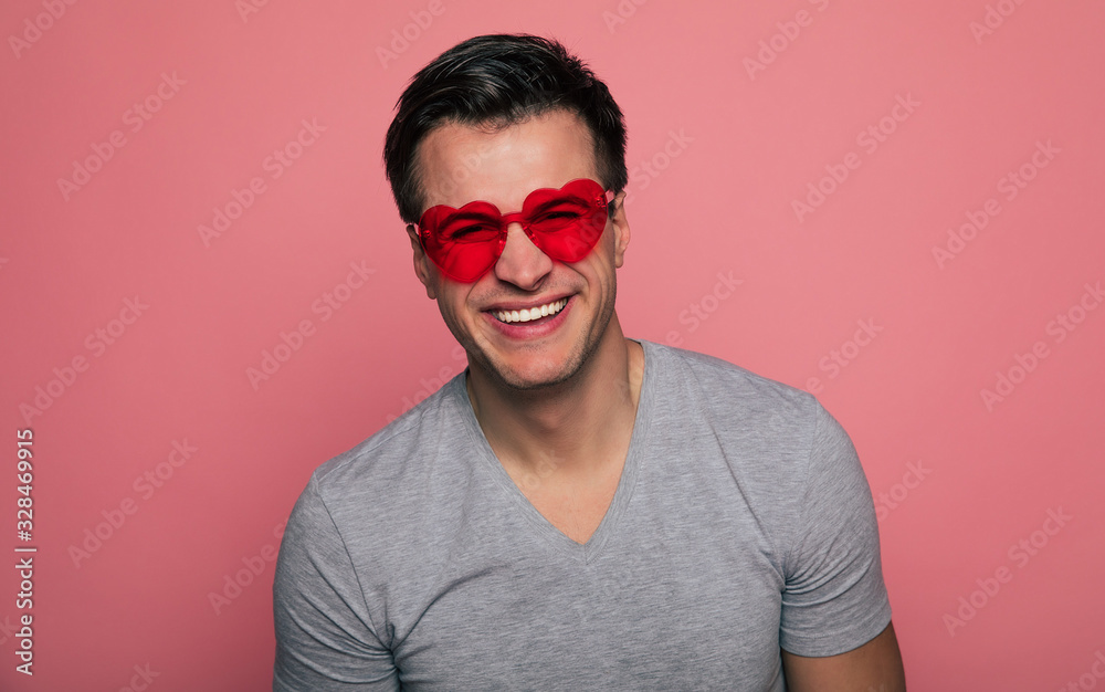 Romantic macho. Valentines day. Happy handsome young man in t-shirt and red glasses in shape of a heart is posing over pink background.
