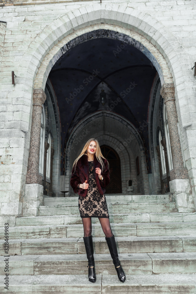 Young girl dressed fur coat and dress standing and look straight on the stairs of church. Old church on the background.