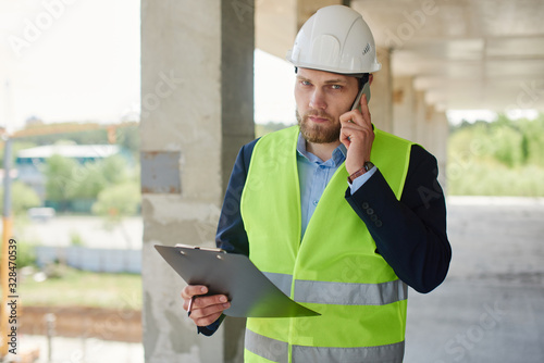 An engineer in a helmet talks on the phone and holds a notepad in a close-up.
