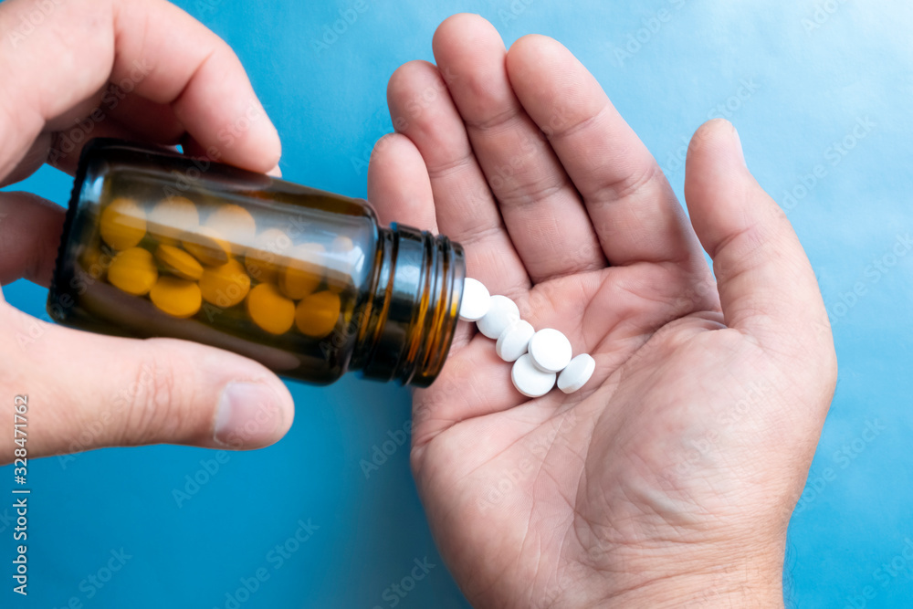 Closeup of man's hand pouring some pills in his hand from a brown bottle. Blue background. Cure concept. Closeup of man's hand pouring some pills in his hand from a brown bottle. Blue background. Cure concept.
