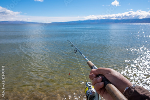 Man's hand is holding a rishing rod with unfocused lake, mountains and sky background.