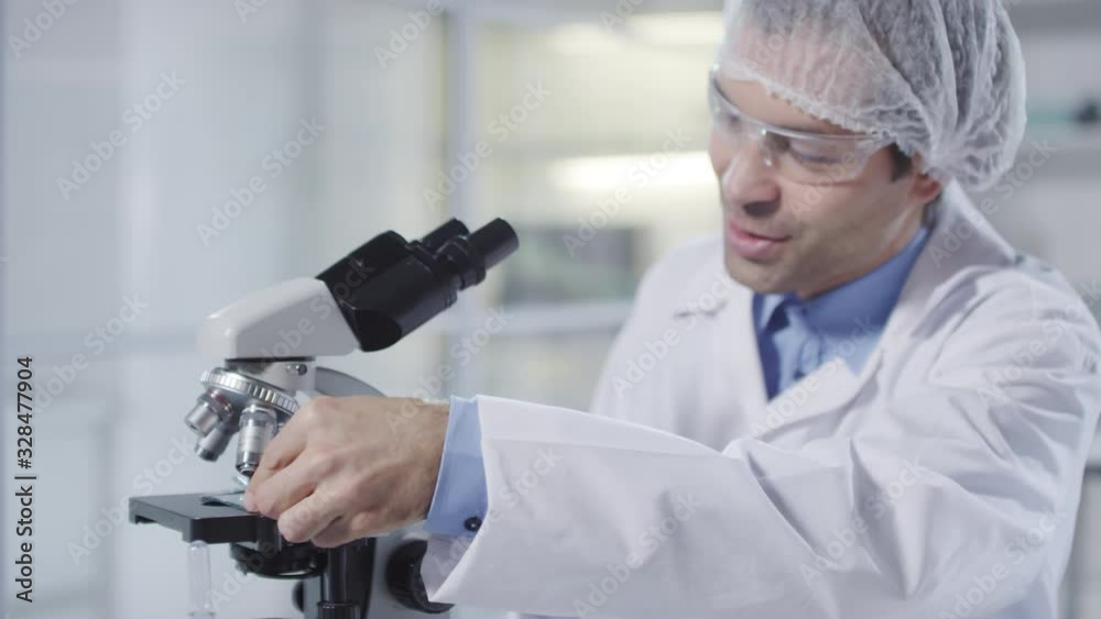 Tracking shot of medical scientist or microbiologist in bouffant cap, lab coat and goggles smiling and looking at sample under microscope Tracking shot of medical scientist or microbiologist in bouffant cap, lab coat and goggles smiling and looking at sample under microscope