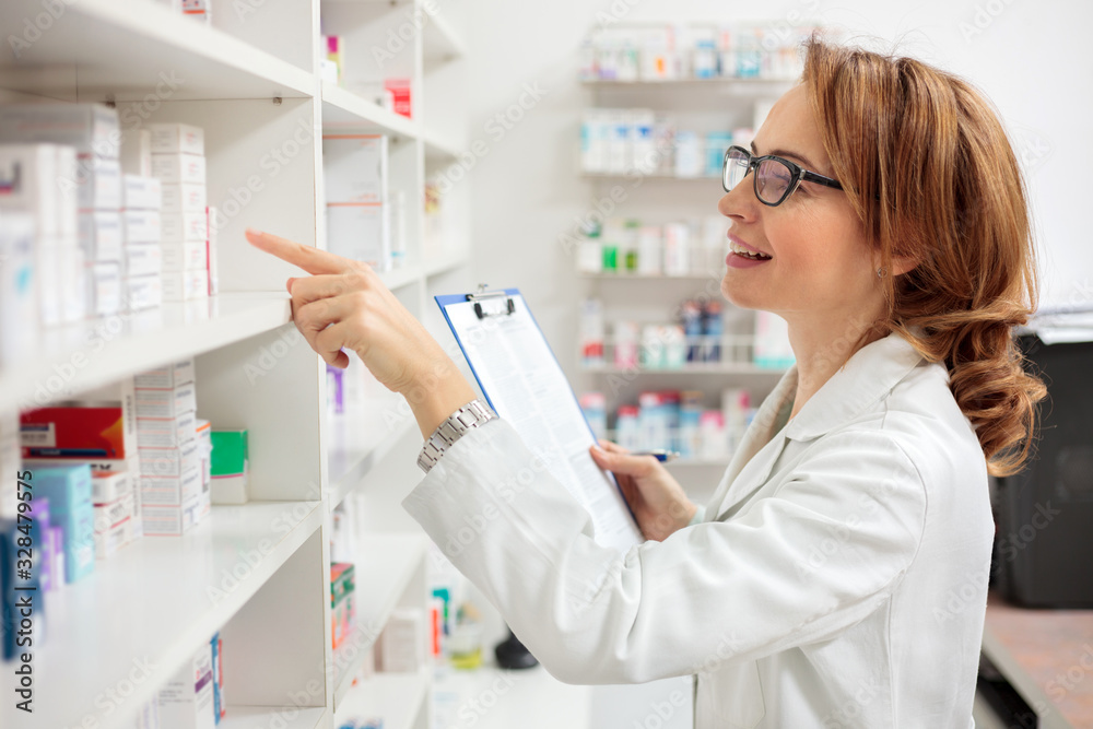 Smiling mid adult female pharmacist holding a clipboard and checking medication boxes on the shelves, working in a pharmacy. Healthcare and medicine concept.