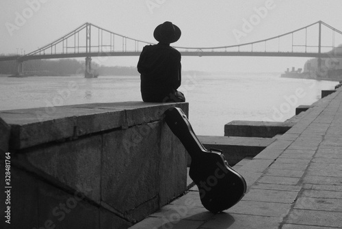 Man in hat and black clothes sits on the river embankment. Near him stands a cover from guitar. Case. Bridge over the river. Back view photo