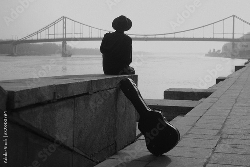 Canvas Print Man in hat and black clothes sits on the river embankment