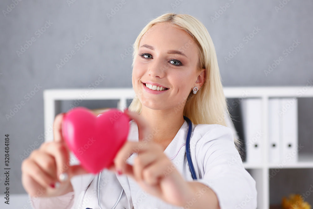 Beautiful smiling blond female doctor hold in arms red toy heart closeup. Cardio therapeutist student education CPR 911 life save physician make cardiac physical pulse rate measure arrhythmia Beautiful smiling blond female doctor hold in arms red toy heart closeup. Cardio therapeutist student education CPR 911 life save physician make cardiac physical pulse rate measure arrhythmia