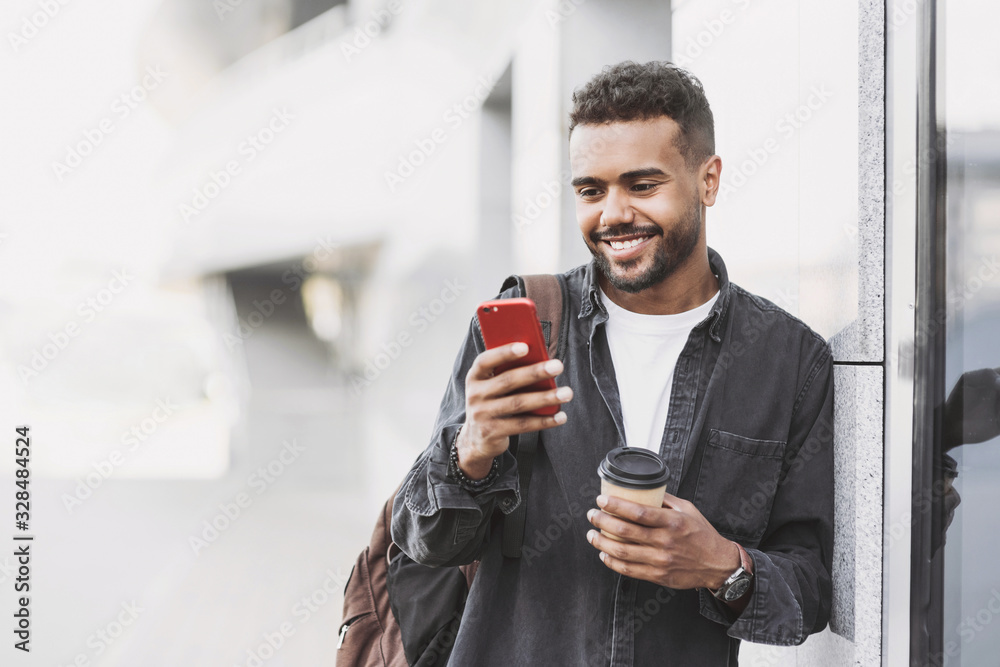 Young handsome men using smartphone in a city. Smiling student man texting on his mobile phone. Coffee break. Modern lifestyle, connection, business concept - obrazy, fototapety, plakaty 