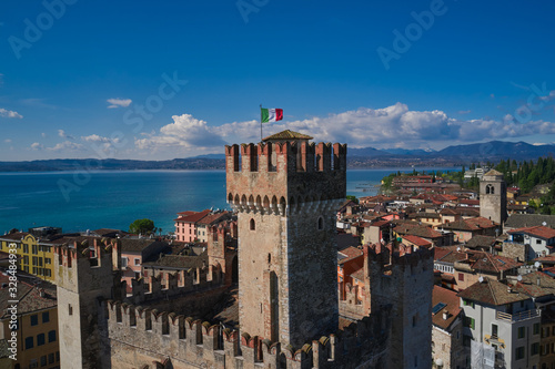 Sirmione castle main tower, close up of Italy flag. In the background mountains in the snow and blue sky. The historical part of the city.