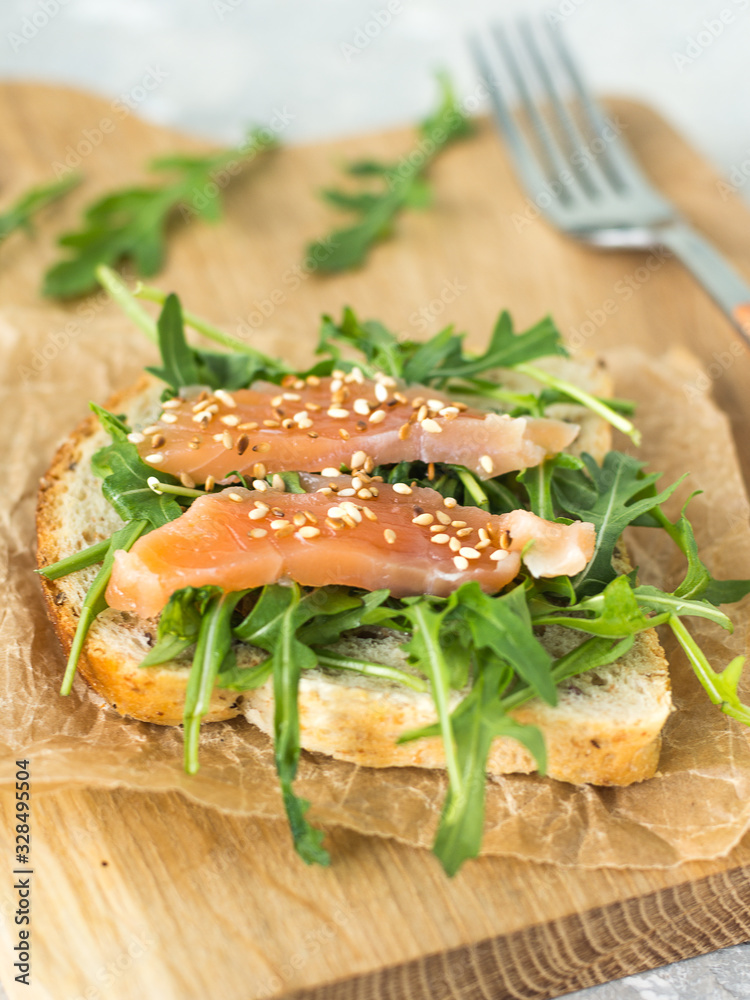 Cereal toast with arugula and trout with sesame seeds on paper and a wooden board top view