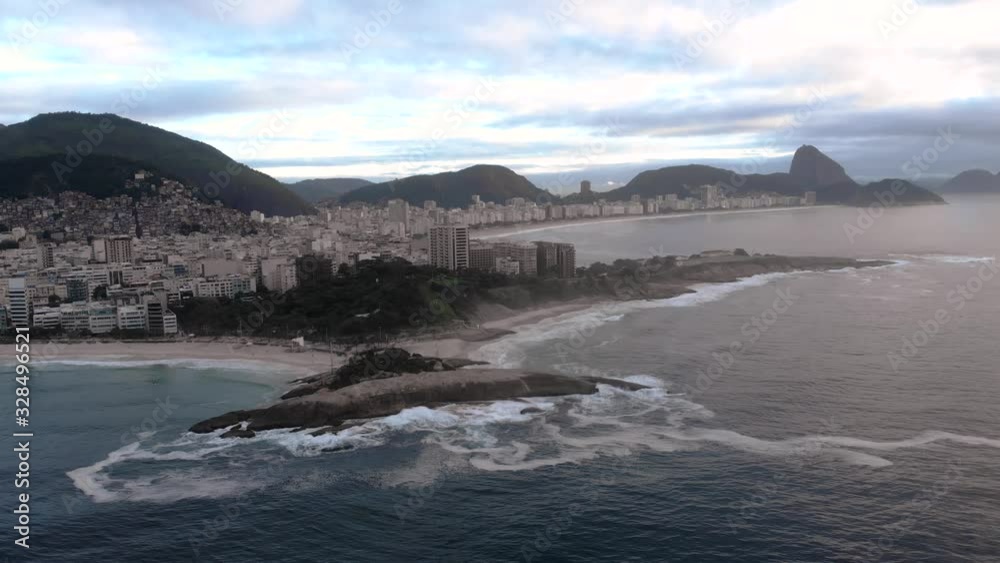 Steady aerial view at sunrise with Arpoador cliff in the foreground with foam created by the incoming crashing waves and Copacabana fort and Sugarloaf mountain in the background Steady aerial view at sunrise with Arpoador cliff in the foreground with foam created by the incoming crashing waves and Copacabana fort and Sugarloaf mountain in the background