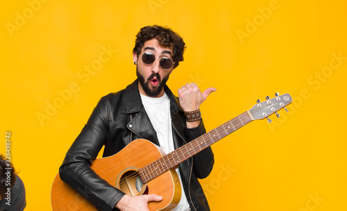 young musician man looking astonished in disbelief, pointing at object on the side and saying wow, unbelievable with a guitar, rock and roll concept photo