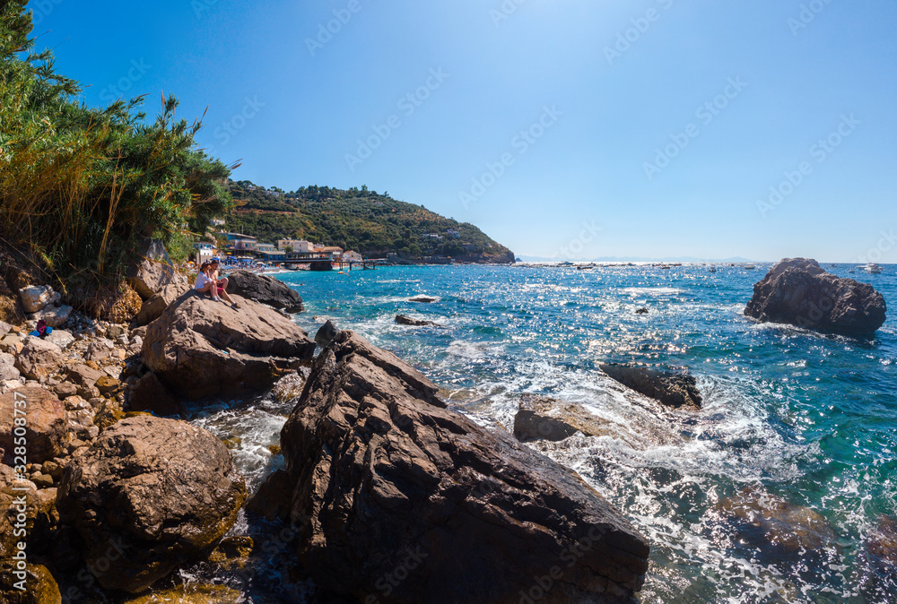 View of paradise, couple of tourists is sitting and look at distace. coastline of the village of Nerano. Wild beach of Italy. Turquoise, blue surface of the water. Vacation and travel concept.