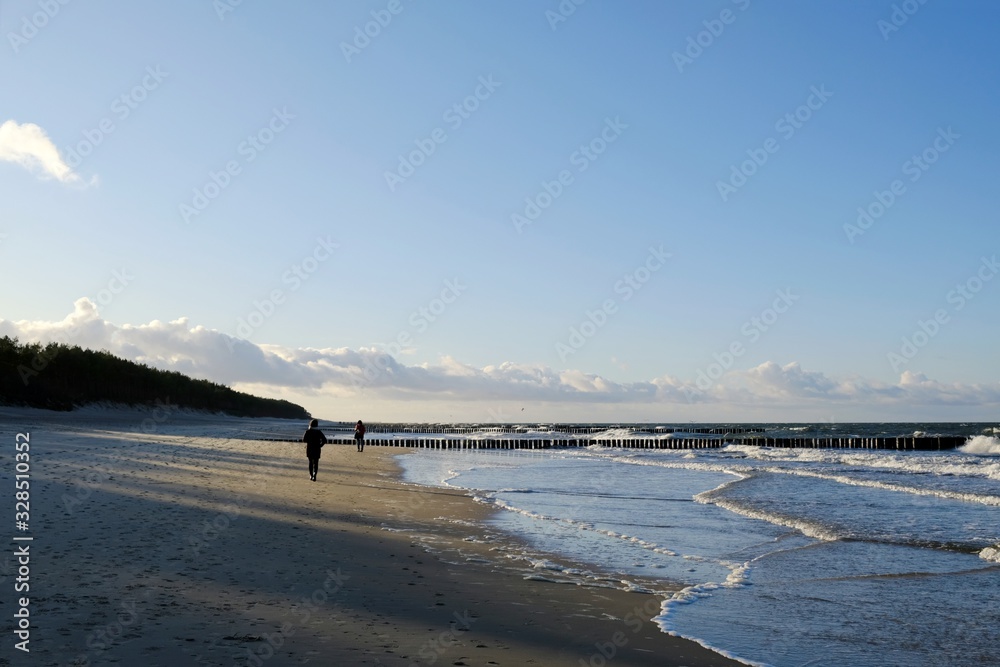 Two persons walking on beach of sea. Waves crashing on the breakwater . Baltic Sea, Dziwnowek, Poland