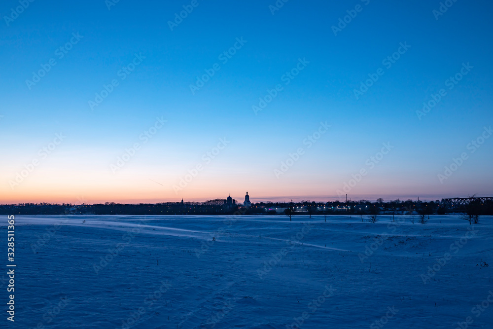 Snowy plain under the sunset sky. Bell tower small town skyline. 