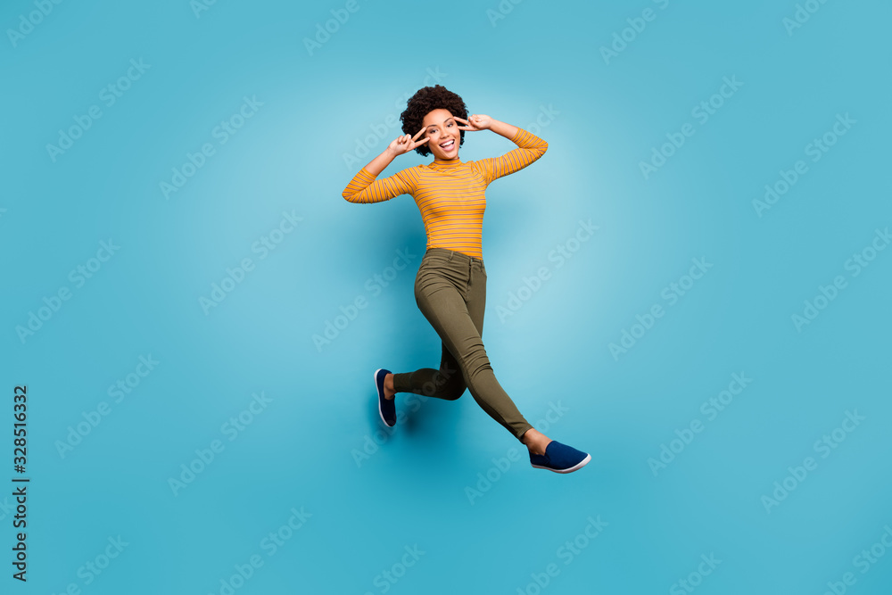 Full size photo of cheerful afro american girl have fall weekends holidays jump make v-signs wear yellow green stylish clothing isolated over blue color background