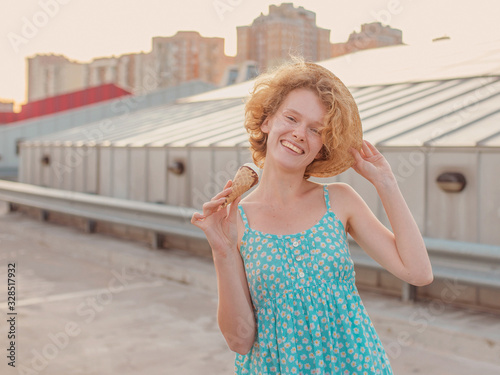 young happy cheerful curly redhead woman in straw hat, blue sundress eating ice cream on skyscraper roof (skyroof). Fun, urban, modern, roof, city, summer, fashion, youth concept photo