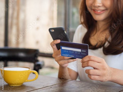 The smiling young Asian woman enjoys shopping online via a smartphone and paying online via credit card. Convenience of spending without cash.