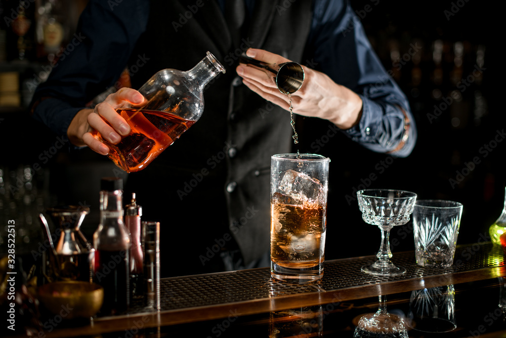 Barman pouring required ingredient for cocktail from bottle with brown alcohol drink.