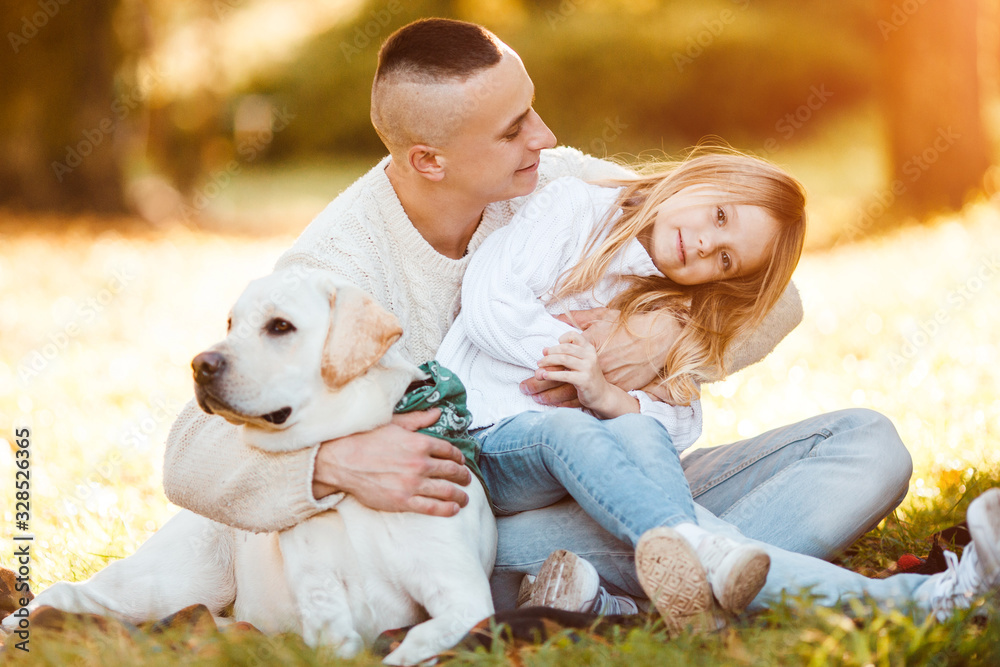 Happy father and daughter with dog labrador is having fun are sitting on green grass in park.