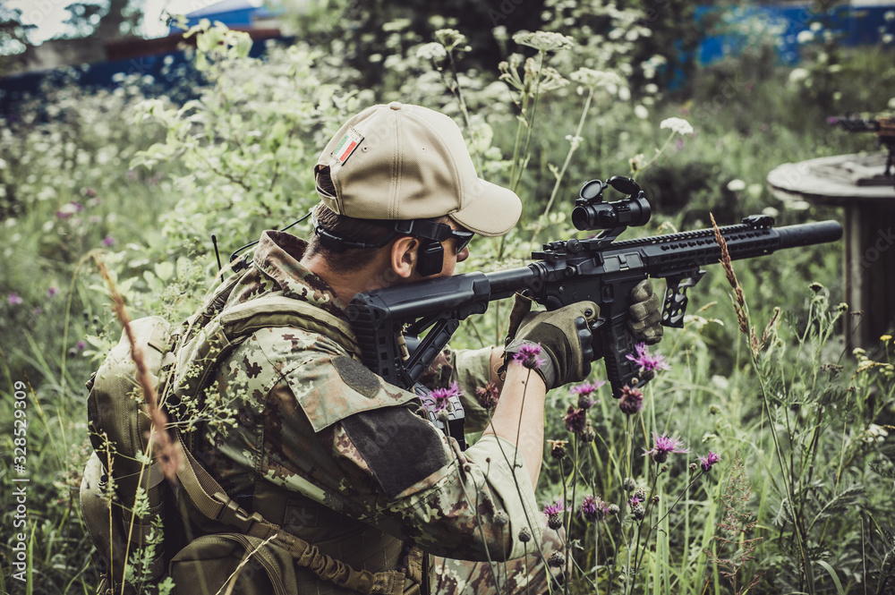 A military man or airsoft player in a camouflage suit sits in the grass and aims from an automatic rifle