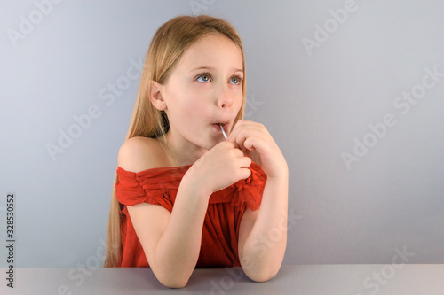 Front view of a beautiful positive little girl joyfully opened her mouth to eat candy on stick on blue background. Sweet tooth kids concept. Nutrition and child health. Copyspace