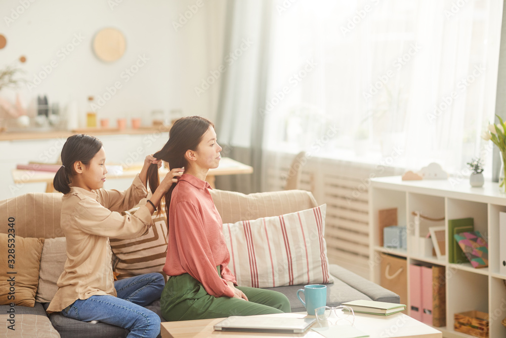 Horizontal side view shot of teen girl sittting on sofa in cozy living room plaiting her mother's hair Horizontal side view shot of teen girl sittting on sofa in cozy living room plaiting her mother's hair