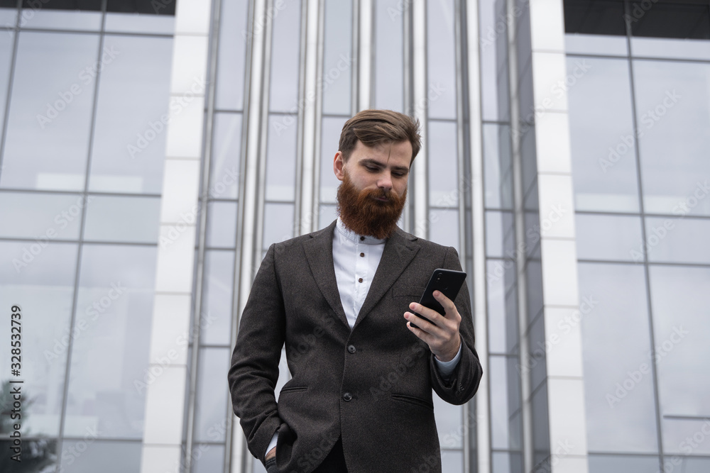 Young bearded Businessman holding mobile smartphone using app texting sms message wearing jacket outdoor. Successful entrepreneur dressed in formal wear communicating with a business partner online. Young bearded Businessman holding mobile smartphone using app texting sms message wearing jacket outdoor. Successful entrepreneur dressed in formal wear communicating with a business partner online.