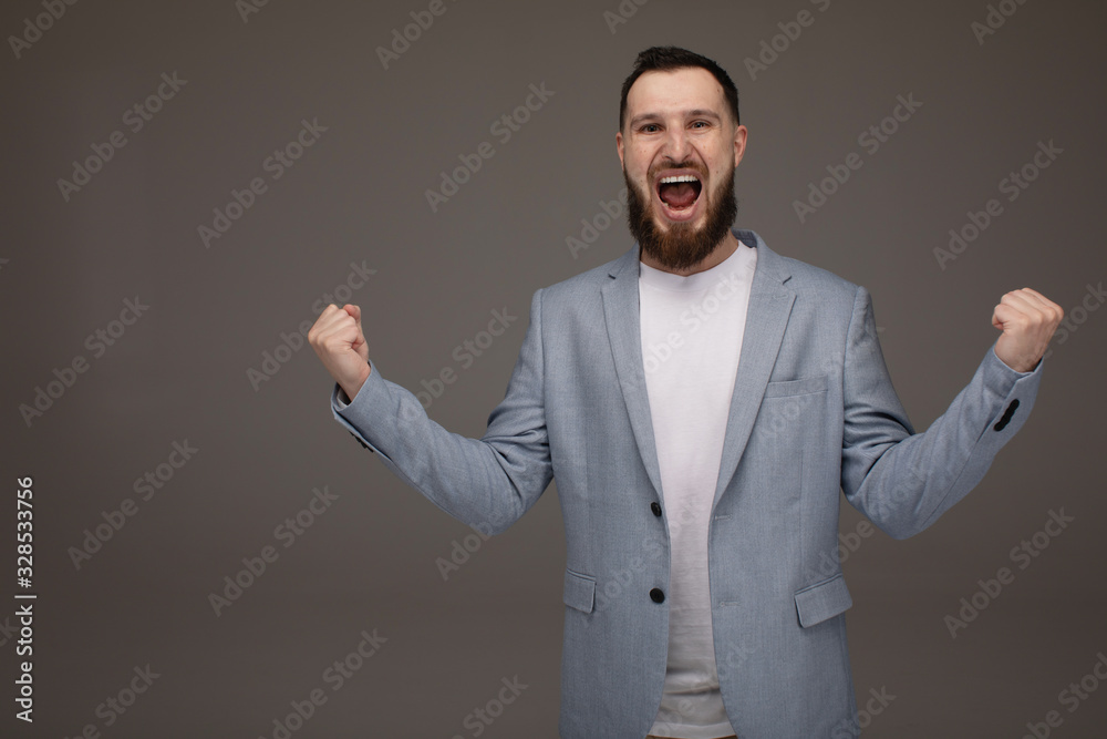 Young handsome bearded business man wearing suit over isolated grey background excited for success with arms raised celebrating victory smiling. Winner concept. Young handsome bearded business man wearing suit over isolated grey background excited for success with arms raised celebrating victory smiling. Winner concept.